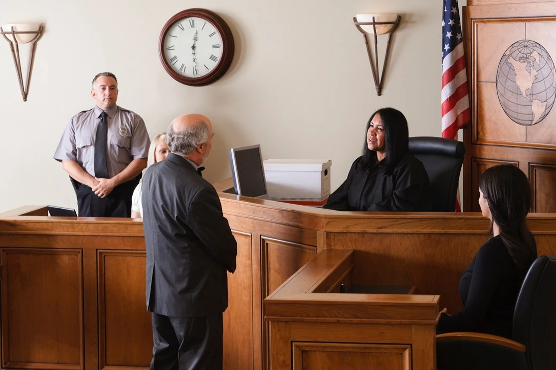 A courtroom scene with a judge seated behind a desk, speaking to a standing lawyer. A security officer stands by, and two women are seated. An American flag and clock are on the wall.