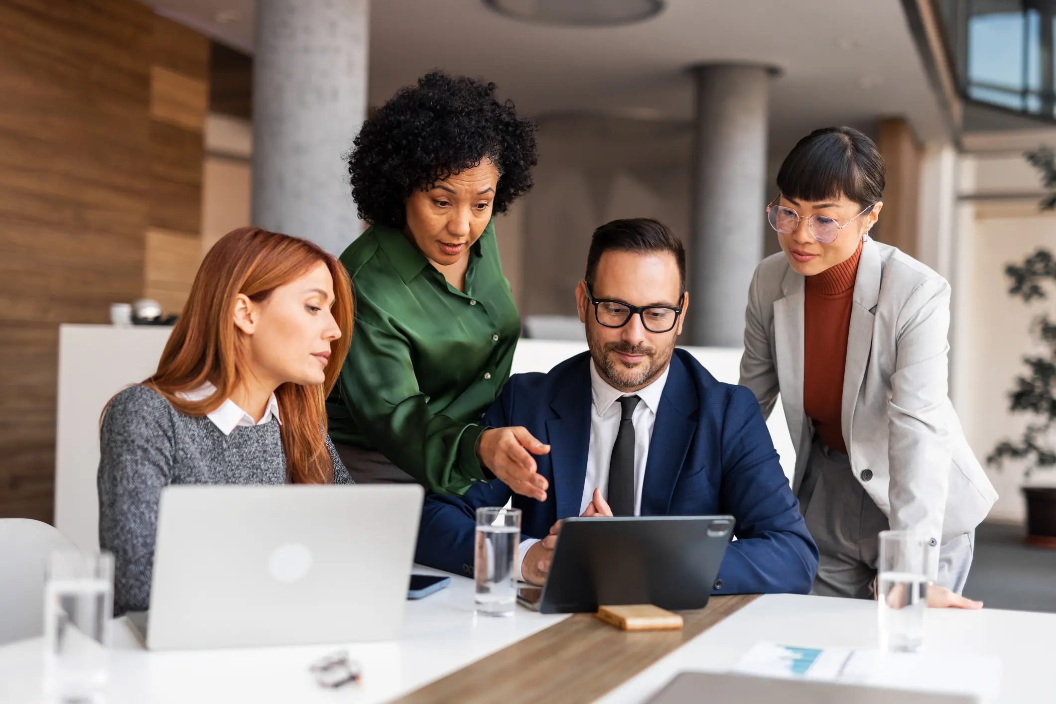 A diverse group of four professionals engage in discussion around a tablet in a modern office setting, conveying collaboration and focus.