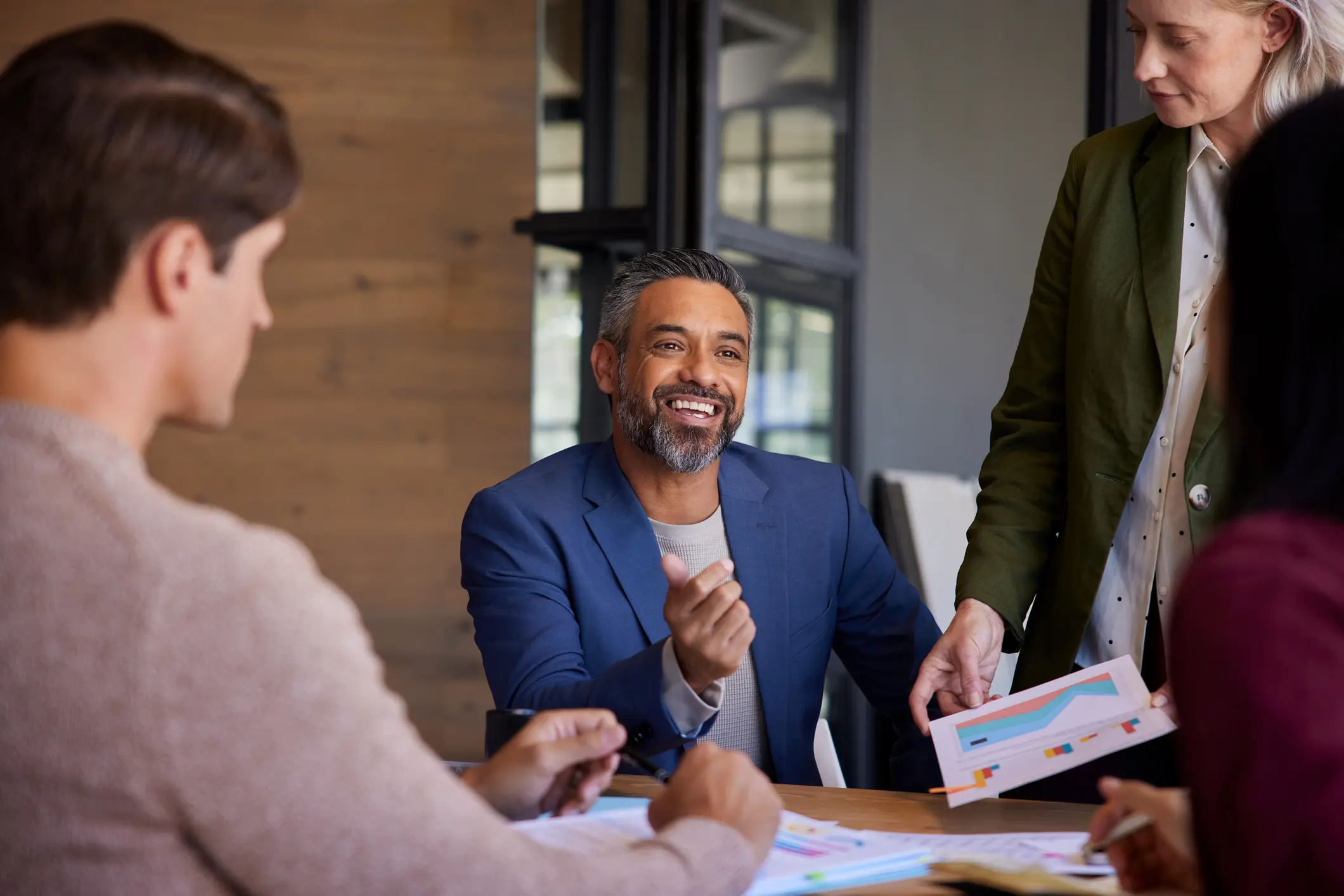 Smiling man in a blue suit gestures during a meeting. Three colleagues, including a woman with papers, engage around a table. Bright, collaborative setting.