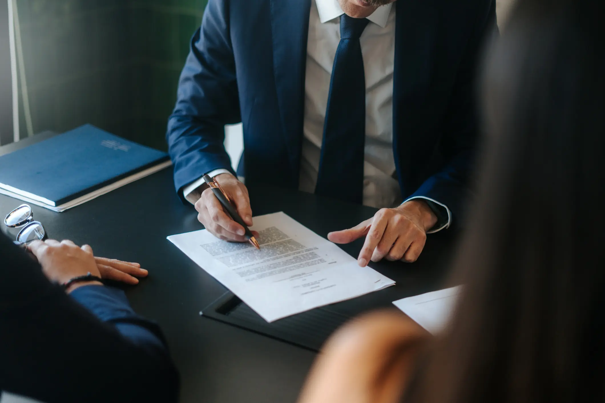 A person in a suit is signing a document at a table, with focused hands and a pen. Two other people are partially visible, creating a formal, business-like atmosphere.