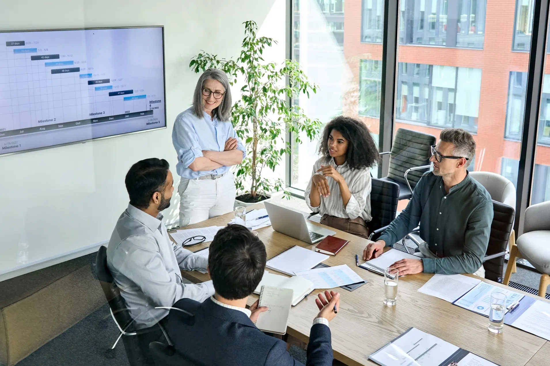BusinessImage A diverse group of five people in a modern office having a meeting. A woman stands by a Gantt chart on a screen, while others sit with laptops and papers, engaged in discussion.