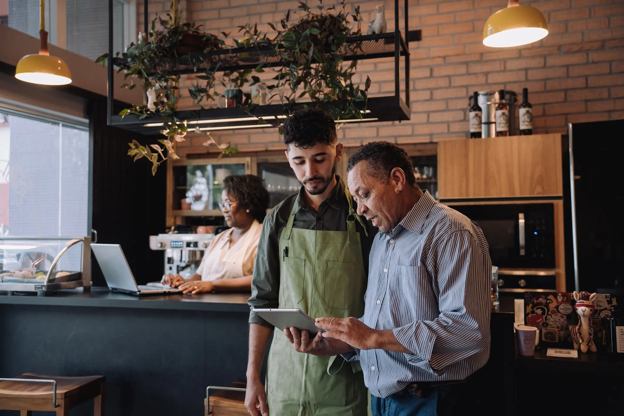 BusinessImage A barista wearing a green apron and a customer view a tablet together in a cozy café. A barista in the background works at an espresso machine.