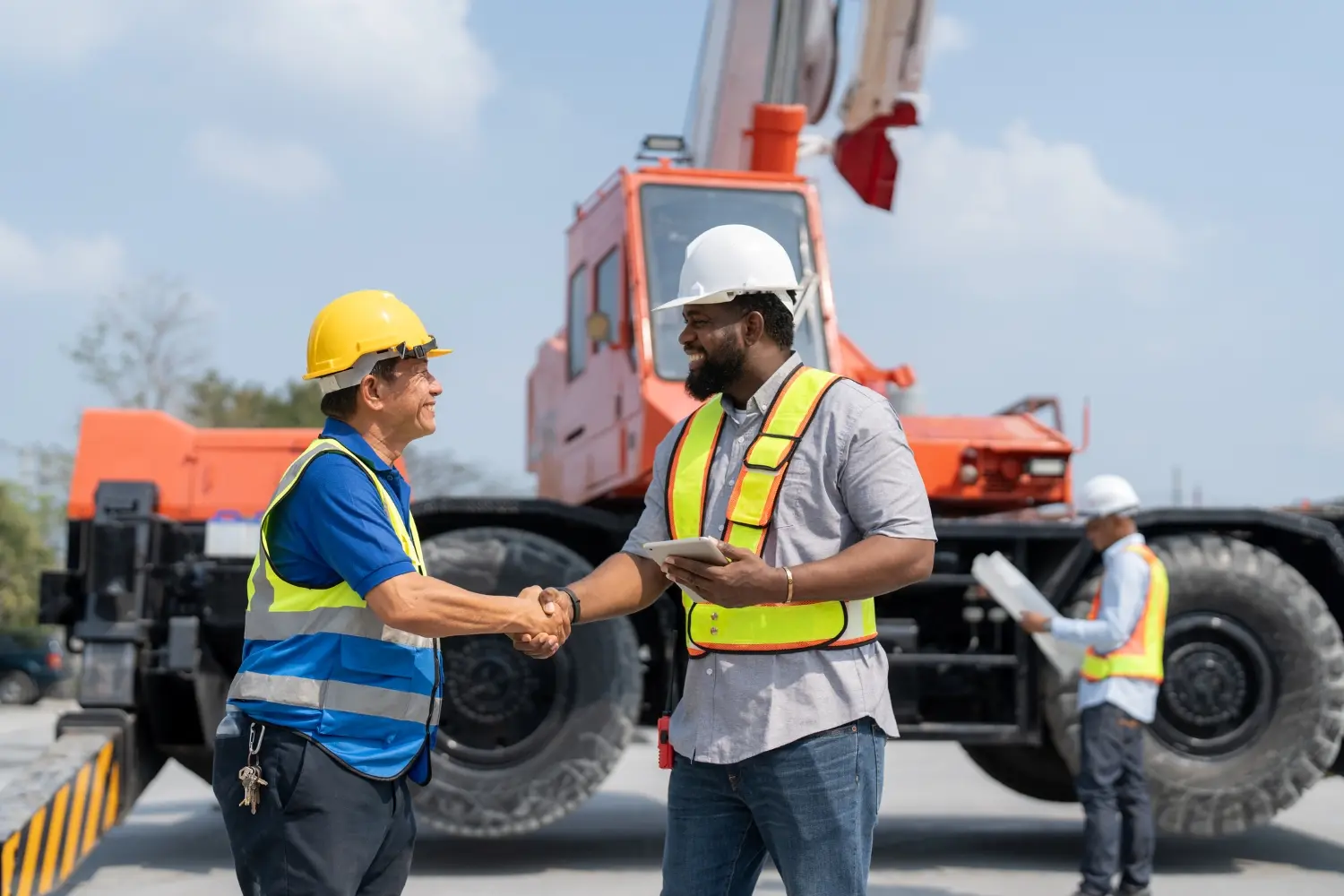 CommercialRealEstateLaw-Hero Two construction workers shake hands, both wearing safety vests and helmets. A crane is in the background, with another worker holding a clipboard. The scene conveys teamwork and collaboration.