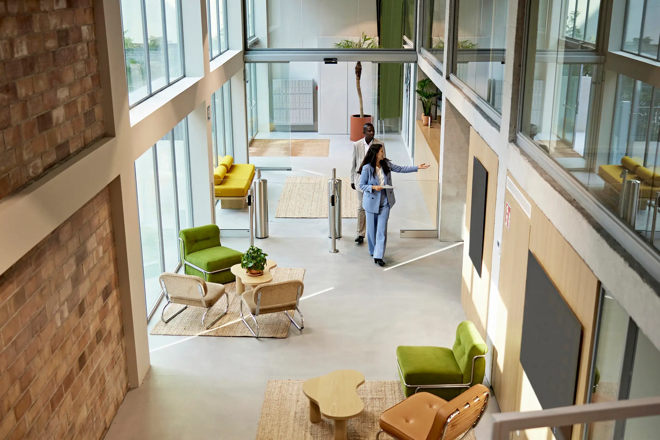 Modern office lobby with large windows and brick walls. Two professionals walk, surrounded by cozy seating, plants, and bright natural light. Calm atmosphere.