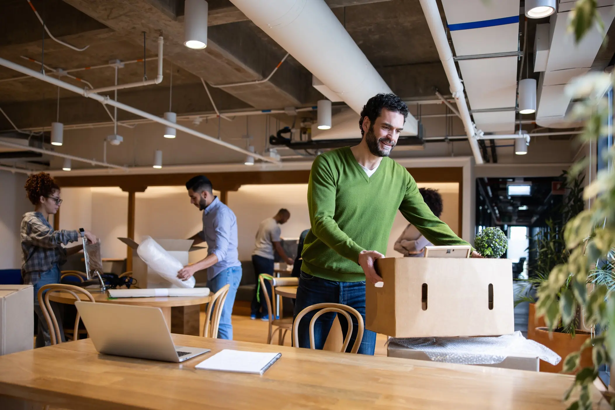 BusinessImage Smiling man in green sweater moving a cardboard box in a modern office. Colleagues in background unpacking and organizing, creating a cheerful atmosphere.