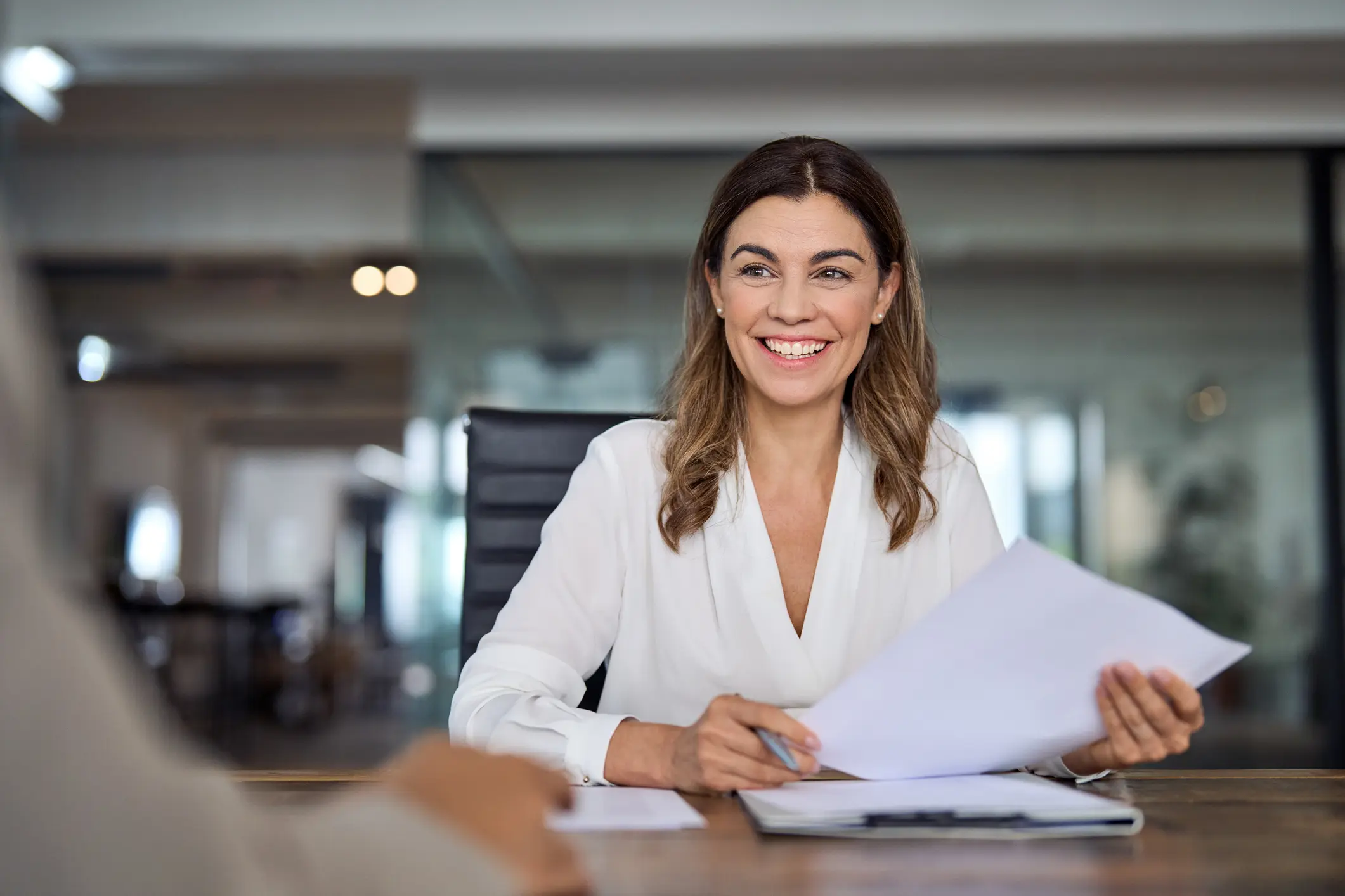 BusinessLigitgation-About Smiling woman in a bright office holding papers, engaging in a conversation. She wears a white blouse, conveying friendliness and professionalism.