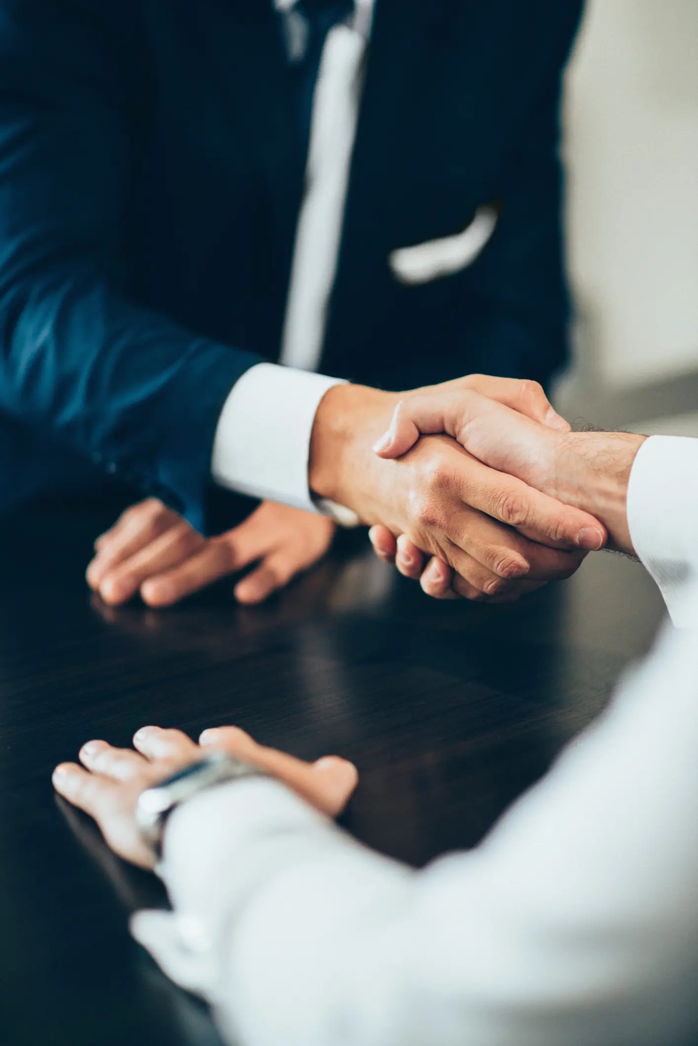 Two people are shaking hands over a dark wooden table. Both wear suits, suggesting a business setting, conveying agreement and professionalism.